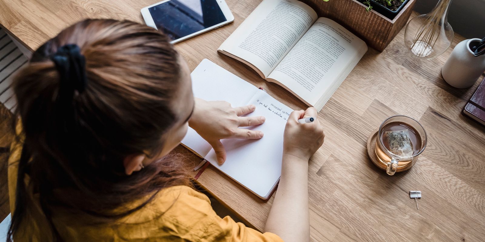 امرأة تكتب في دفتر على مكتب مرتب إلى جانب الكتب وكوب شاي